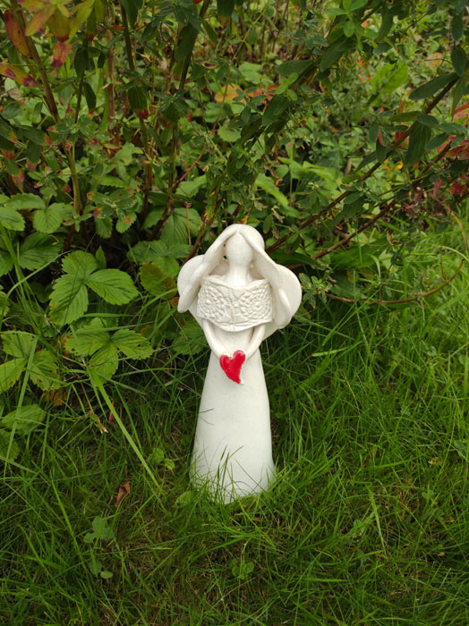Keramický anděl bílý, zdobený krajkou, držící červené srdíčko.